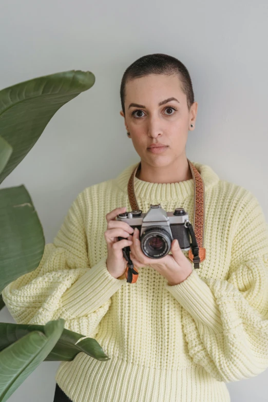 a woman holding a camera in front of a plant, wearing a sweater, portrait sophie mudd, with yellow cloths, jordan lamarre - wan