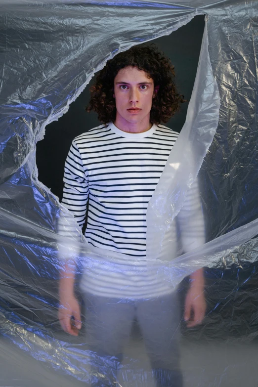 a man standing in a room covered in plastic, an album cover, inspired by Christo, unsplash, curly haired, wearing stripe shirt, containment pod, with a black background