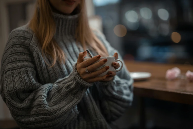 a woman in a sweater holding a cup of coffee, by Adam Marczyński, trending on pexels, aestheticism, mysterious coffee shop girl, soft blur, grey, 8k 50mm iso 10