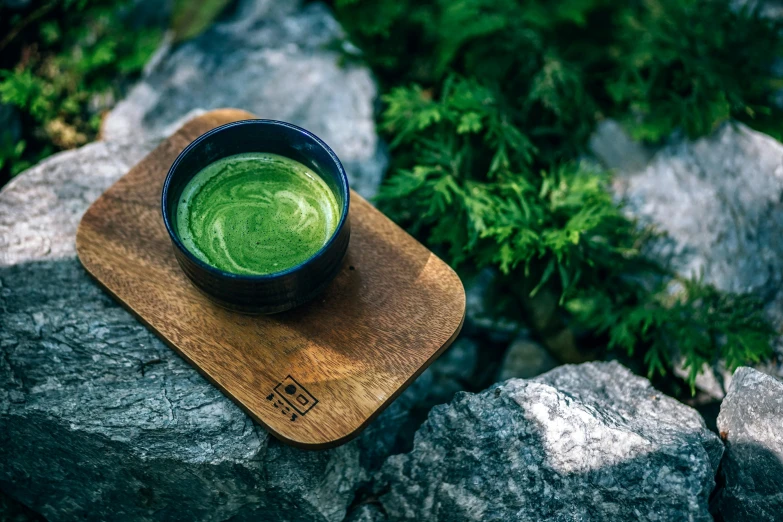 a bowl of green tea sitting on top of a wooden coaster, inspired by Kanō Shōsenin, trending on unsplash, hurufiyya, outdoors, moringa juice, shot on hasselblad, cliffside