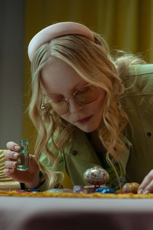 a woman sitting at a table in front of a cake, wizard examining eggs, from netflix's arcane, pale skin curly blond hair, yellow carrera glasses