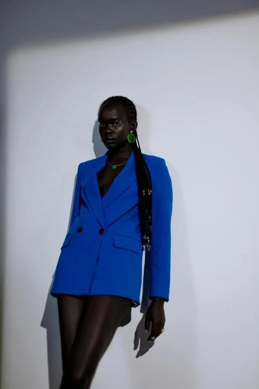 a woman in a blue suit standing next to a white wall, by Olivia Peguero, adut akech, short skirt and a long jacket, dayglo blue, thumbnail