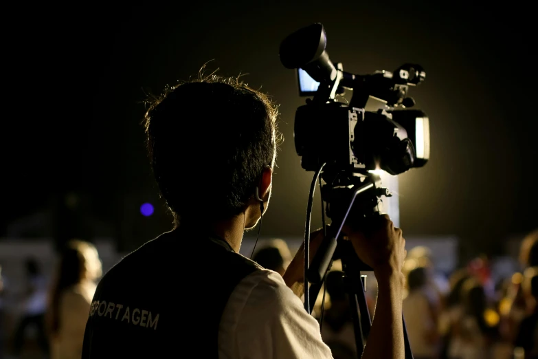 a man holding a camera in front of a crowd, spotlights, cinematográfica, back facing the camera, journalism