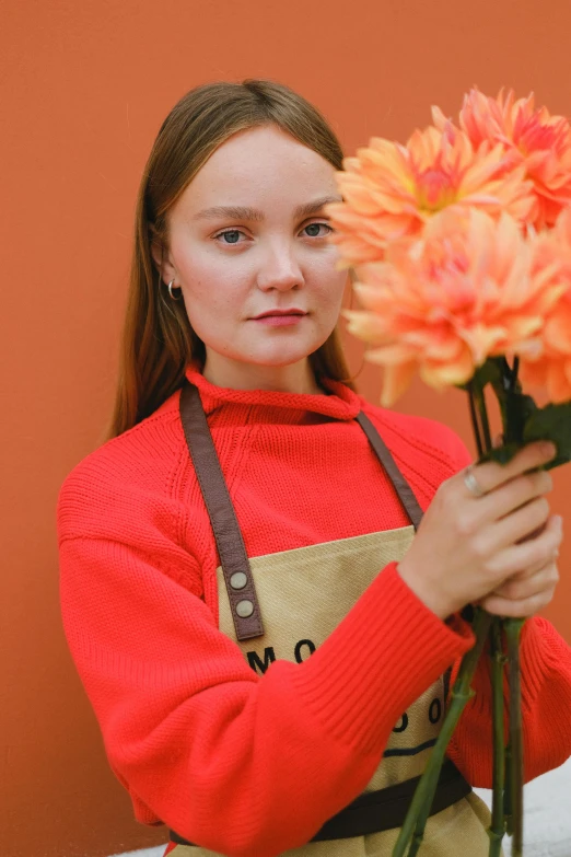 a woman in an apron holding a bunch of flowers, by Julia Pishtar, pexels contest winner, magic realism, long orange sweatshirt, dakota fanning, teenager girl, product introduction photo