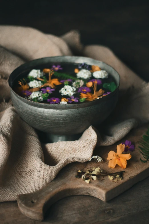 a bowl filled with flowers sitting on top of a wooden cutting board, by Else Alfelt, trending on unsplash, black oil bath, medieval feast, dark grey and orange colours, made of flowers and leaves