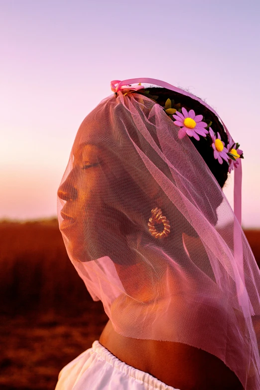 a woman standing in a field with a veil over her head, inspired by Carrie Mae Weems, trending on pexels, wearing a flower headpiece, pink sunset hue, glowing details, doja cat