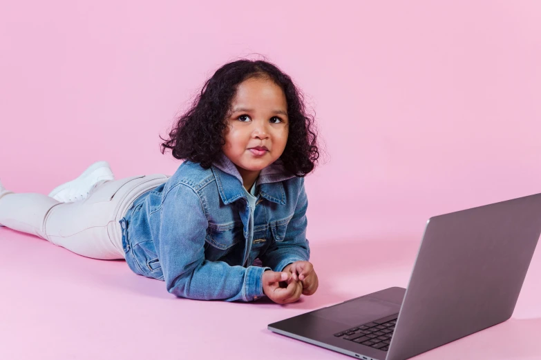 a little girl laying on the floor next to a laptop, trending on pexels, computer art, indian girl with brown skin, pink hue, denim, looking straight to camera