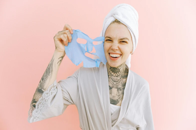 a woman with a towel on her head holding a piece of paper in front of her face, an album cover, pexels contest winner, antipodeans, smiling mask, freezing blue skin, tattooed body, skincare