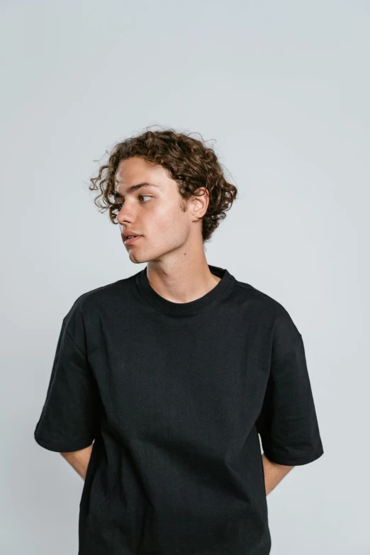 a young man with curly hair wearing a black t - shirt, trending on unsplash, plain background, 1 6 years old, baggy, shorter neck