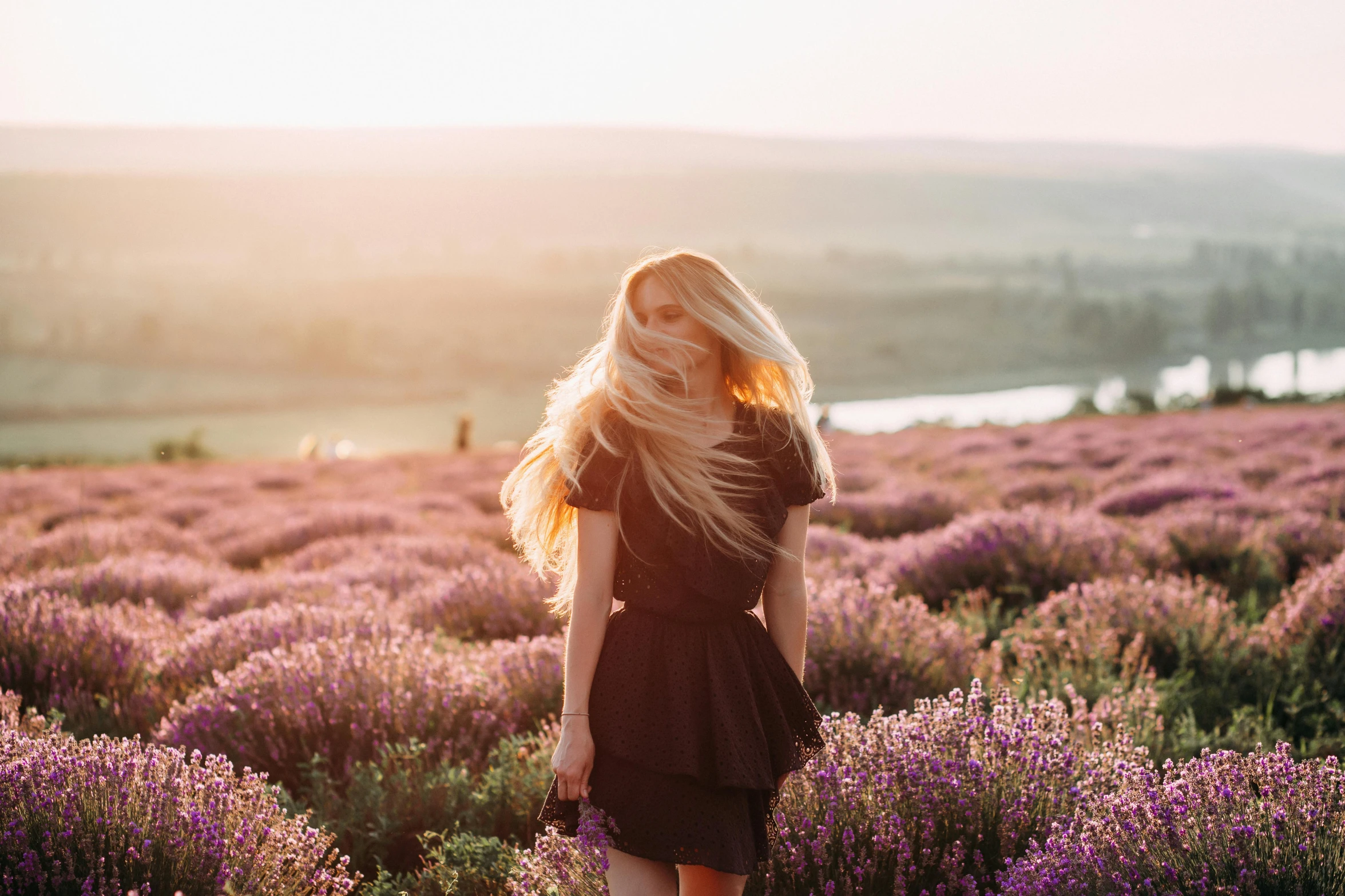 a woman standing in a field of purple flowers, pexels contest winner, flowing blonde hair, in a sunset haze, photoshoot for skincare brand, wearing a dark dress