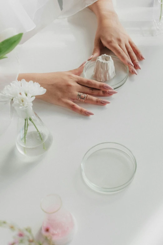 a woman sitting at a table with a vase of flowers, trending on pexels, partially cupping her hands, cosmetics, white details, clear liquid