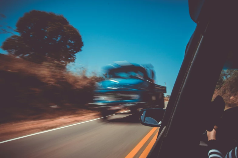 a blue truck driving down a road next to a forest, a picture, unsplash, photorealism, high speed chase, hotrods driving down a street, thumbnail, low - angle shot