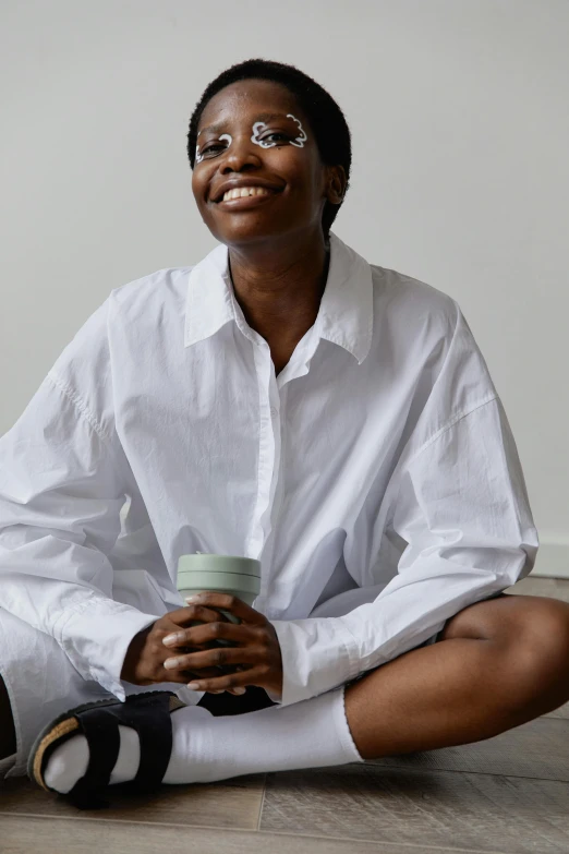 a woman sitting on the floor holding a cup, by Nina Hamnett, trending on unsplash, white wrinkled shirt, dark-skinned, smiling, lab coat