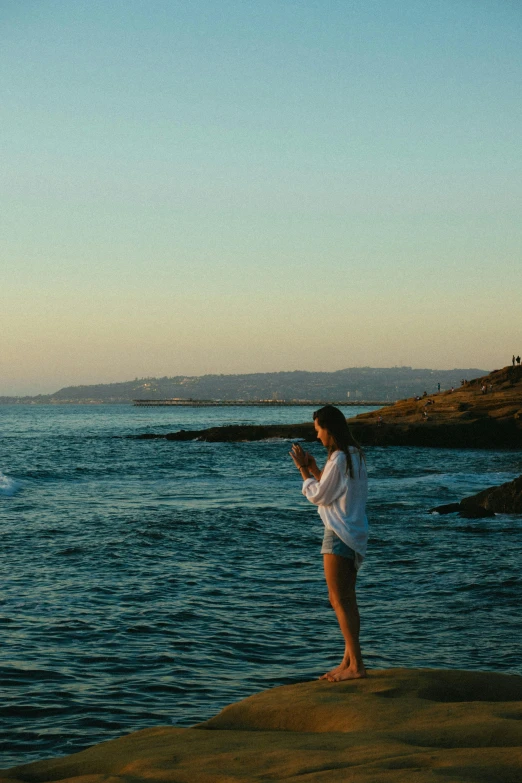 a woman standing on top of a rock next to the ocean, in the evening, people angling at the edge, lorde, with a long white