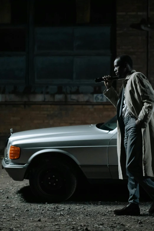 a man in a trench coat standing next to a car, inspired by Gordon Parks, pexels contest winner, dark skinned, movie still 8 k, with a gun, grey skinned