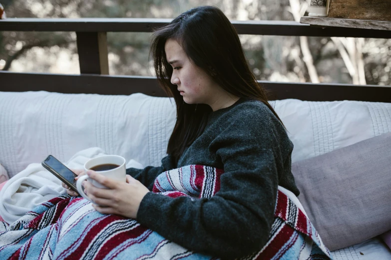 a woman sitting on a couch holding a cup of coffee, trending on pexels, happening, covered with blanket, outside, asian female, gif