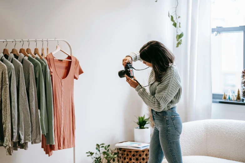 a woman standing in front of a clothing rack holding a camera, plant photography, inspect in inventory image, cute photograph, green and brown clothes