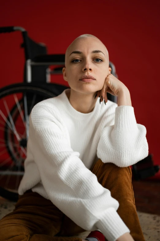 a woman sitting on the floor with a wheelchair in the background, a character portrait, trending on pexels, antipodeans, shaved bald head, white sweater, motorcycle, portrait sophie mudd