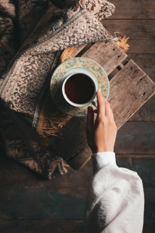 a person holding a cup of tea next to a pile of books, a portrait, inspired by Elsa Bleda, trending on pexels, renaissance, over vivid dark wood table, long sleeves, maroon, gif