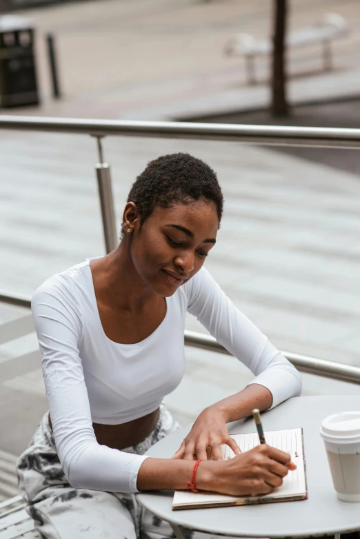 a woman sitting at a table writing on a notebook, by Lily Delissa Joseph, trending on unsplash, academic art, beautiful city black woman only, wearing white v - neck top, high view, profile pose