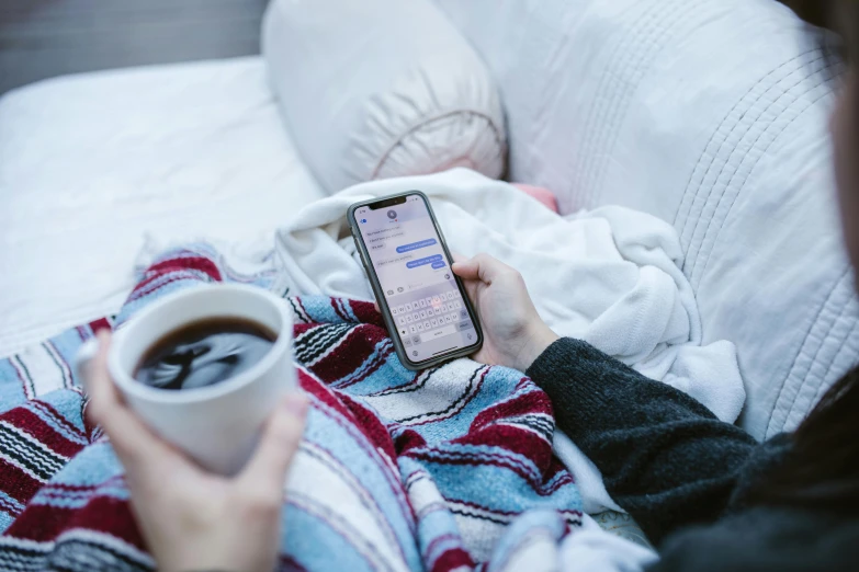 a person laying on a bed holding a cup of coffee, trending on pexels, happening, checking her phone, avatar image, cold freezing nights, nursing