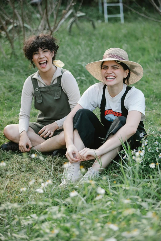 a couple of women sitting on top of a lush green field, an album cover, pexels, wearing a straw hat and overalls, declan mckenna, laughing, julia fuentes
