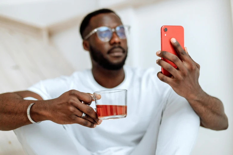 a man in a white shirt holding a red cell phone, a photo, trending on pexels, happening, is ((drinking a cup of tea)), man is with black skin, wearing a red outfit, avatar image