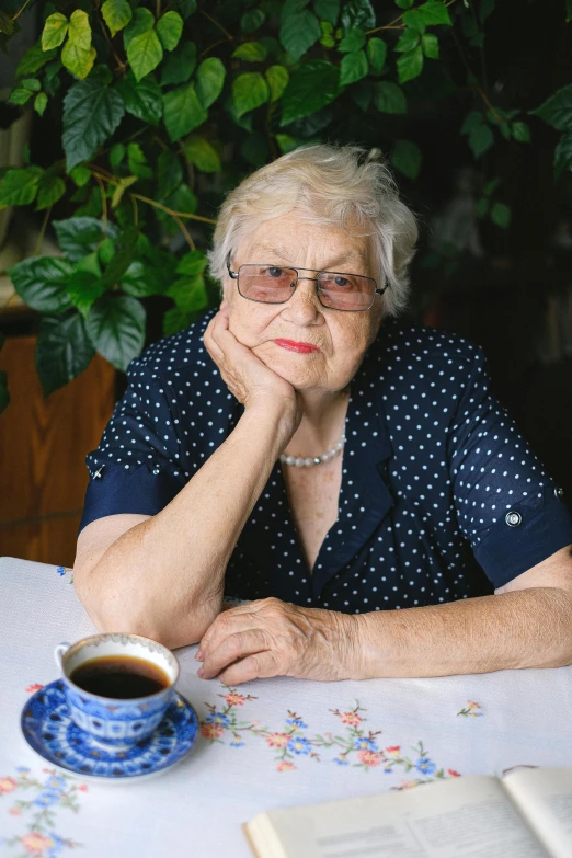 a woman sitting at a table with a cup of coffee, a portrait, by Elsie Few, pexels contest winner, grumpy [ old ], olga zakharova, wearing small round glasses, non-binary