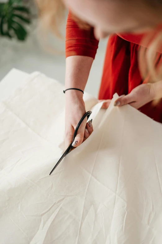 a woman cutting a piece of paper with a pair of scissors, by Haukur Halldórsson, trending on pexels, arts and crafts movement, folds of fabric, where a large, with a square, cream paper