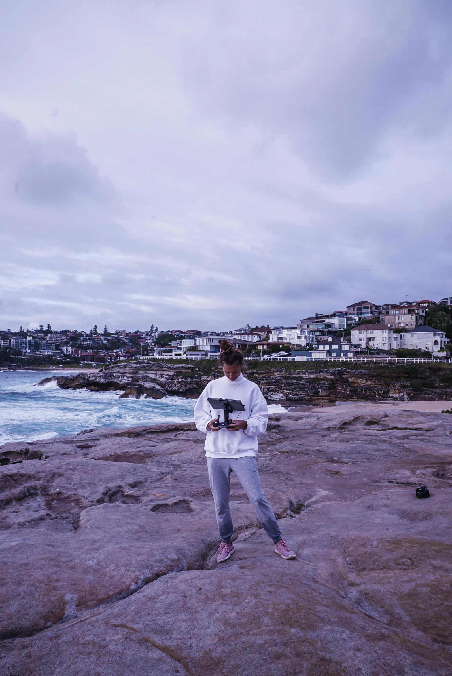 a man standing on top of a rock next to the ocean, a polaroid photo, inspired by Sydney Carline, unsplash, wearing a track suit, purple roofs, bondi beach in the background, panoramic view of girl