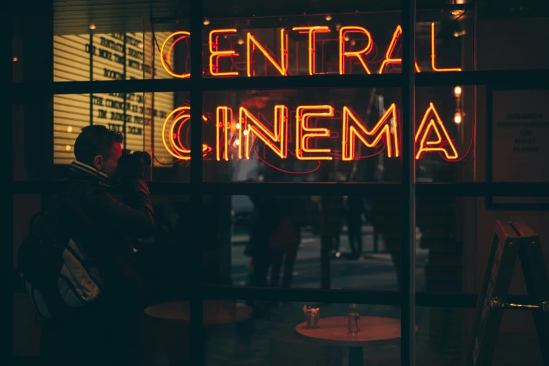 a man standing in front of a sign that says central cinema, by Emma Andijewska, unsplash contest winner, red neon lights inside it, instagram post, cctv footage of a movie set, sitting in a movie theater