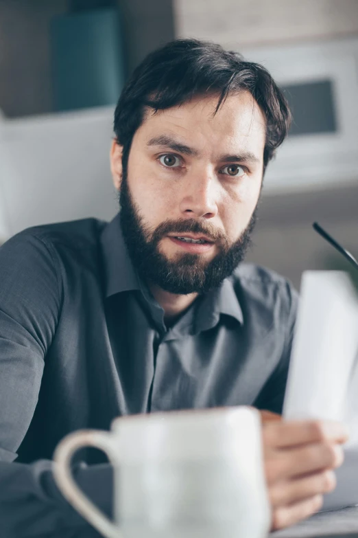 a man sitting at a table with a cup of coffee, scruffy facial hair, pen and paper, grey, annoyed