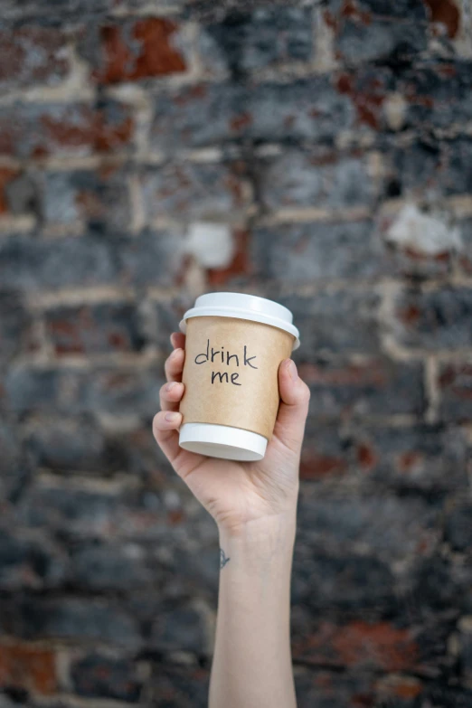 a person holding a cup of coffee in front of a brick wall, by Adam Marczyński, trending on unsplash, label, made of drink, paper cup, light tan