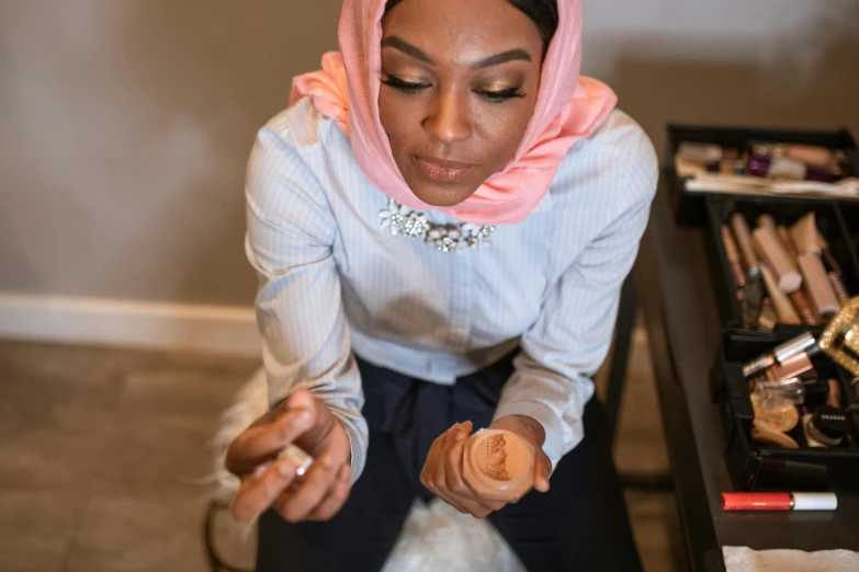 a woman sitting on the floor holding a jar of makeup, inspired by Maryam Hashemi, pexels contest winner, hurufiyya, partially cupping her hands, on a desk, head scarf, ( ( dark skin ) )
