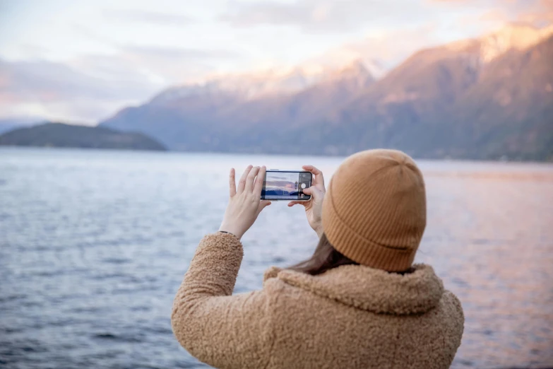 a woman taking a picture with her cell phone, a picture, trending on pexels, fjords in background, warm coloured, product shot, back to camera