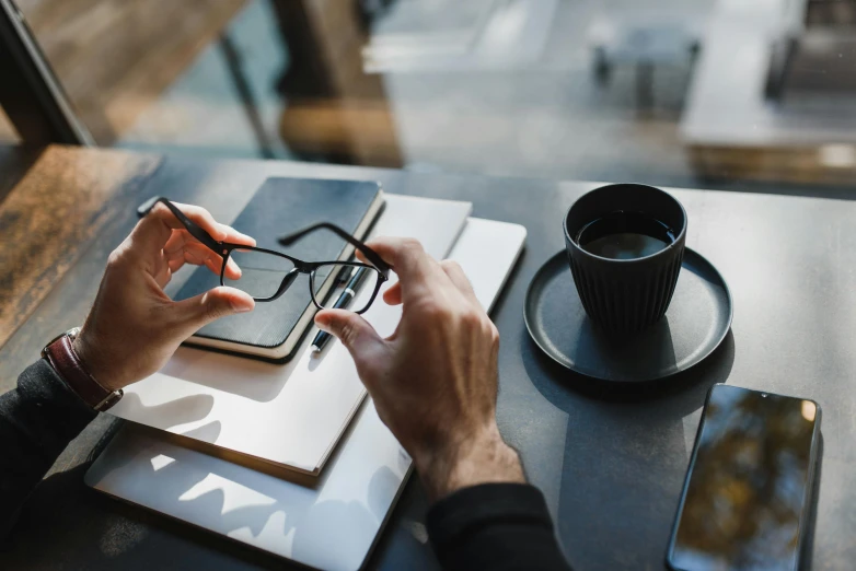 a person sitting at a table with a laptop and a cup of coffee, by Sebastian Vrancx, trending on pexels, wearing black rimmed glasses, holds a smart phone in one hand, maintenance, thumbnail