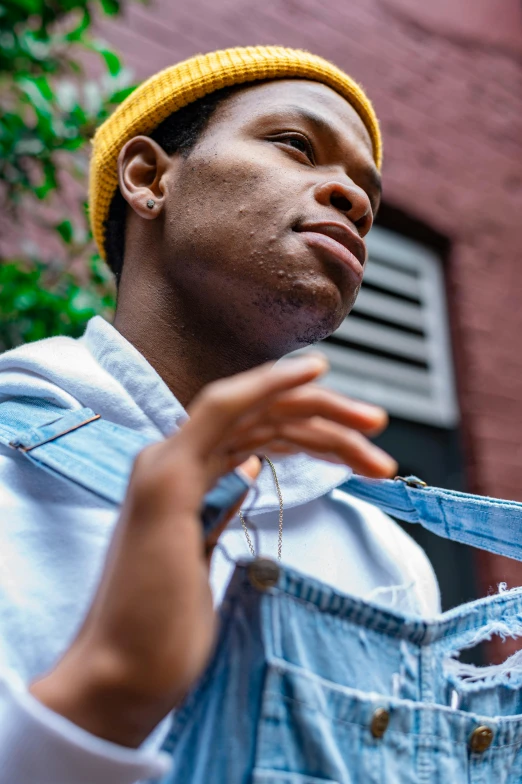 a man standing in front of a brick building, an album cover, by Stokely Webster, trending on pexels, realism, non binary model, wearing blue jean overalls, scratching head, on a yellow canva