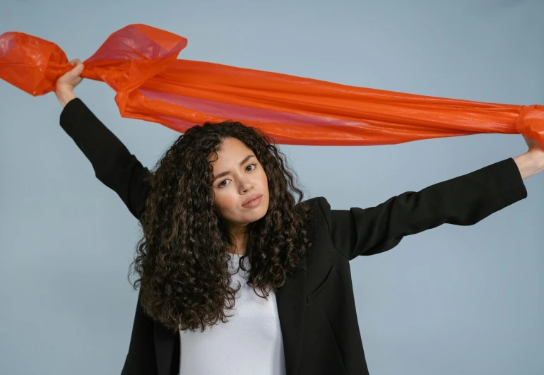 a woman holding a red scarf over her head, an album cover, inspired by Christo, pexels contest winner, antipodeans, tessa thompson, orange neon, future inflatable jacket, power pose