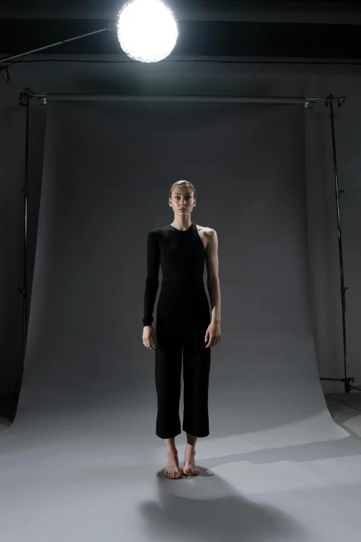 a woman standing in front of a light in a studio, a digital rendering, inspired by Vanessa Beecroft, unsplash, wearing a black catsuit, assymetrical, avant designer uniform, at a fashion shoot