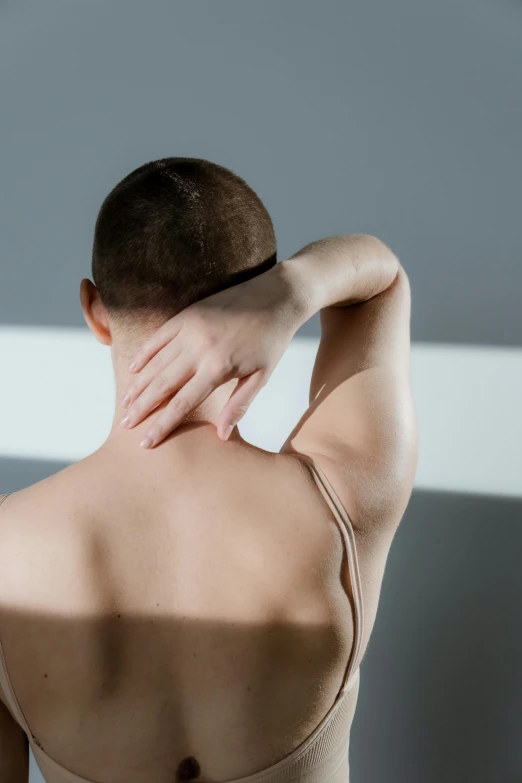 a woman standing in a room with her back to the camera, by Anna Füssli, trending on pexels, renaissance, muscular neck, morning light showing injuries, greeting hand on head, plain background