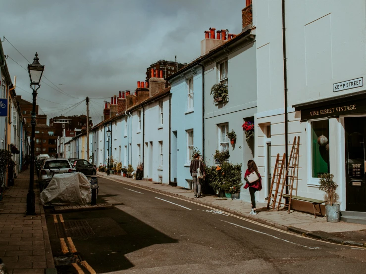 a woman walking down a street past a row of houses, by Rachel Reckitt, pexels contest winner, street of teal stone, cars parked underneath, gif, regency-era