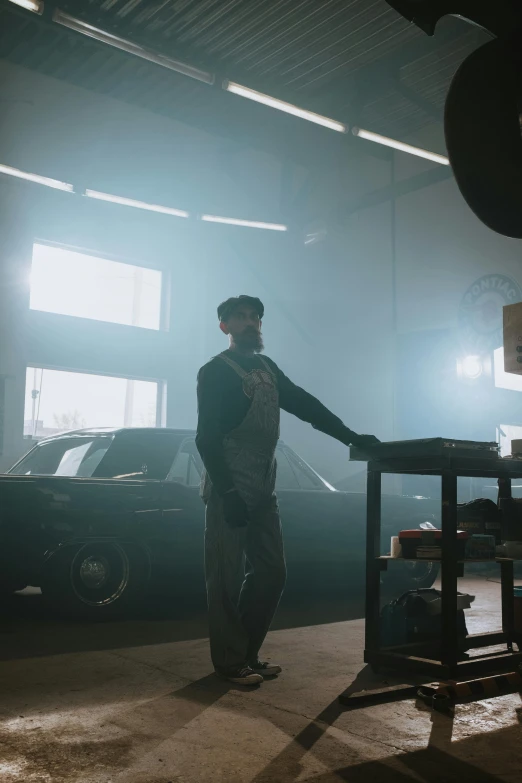 a man standing next to a table in a garage, pexels contest winner, zack snyder cinematography style, smokey atmosphere, mechanic, promo image