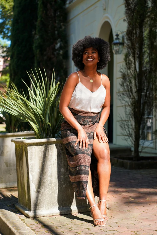 a woman sitting on a bench next to a potted plant, brown skin, wearing a tanktop and skirt, curly haired, sydney park