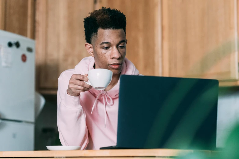 a man sitting at a table with a laptop and a cup of coffee, by Carey Morris, pexels, renaissance, wearing a pink hoodie, avatar image, black teenage boy, drinking tea