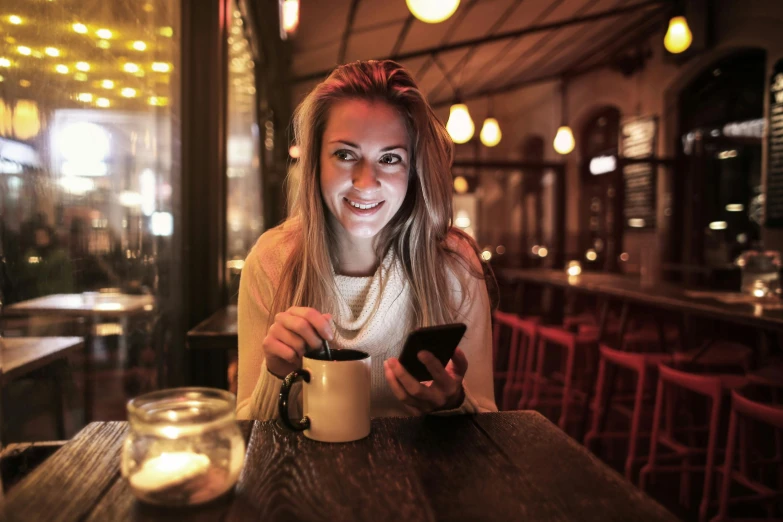 a woman sitting at a table with a cup of coffee, pexels contest winner, happening, night life, holding a very advance phone, avatar image, good looking