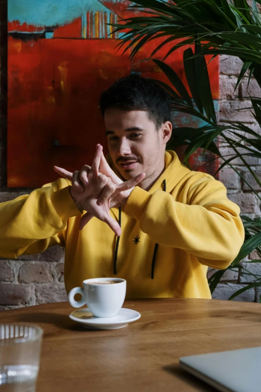 a man sitting at a table in front of a laptop, inspired by Ding Guanpeng, realism, wearing a yellow hoodie, serpentine pose gesture, cup of coffee, gif