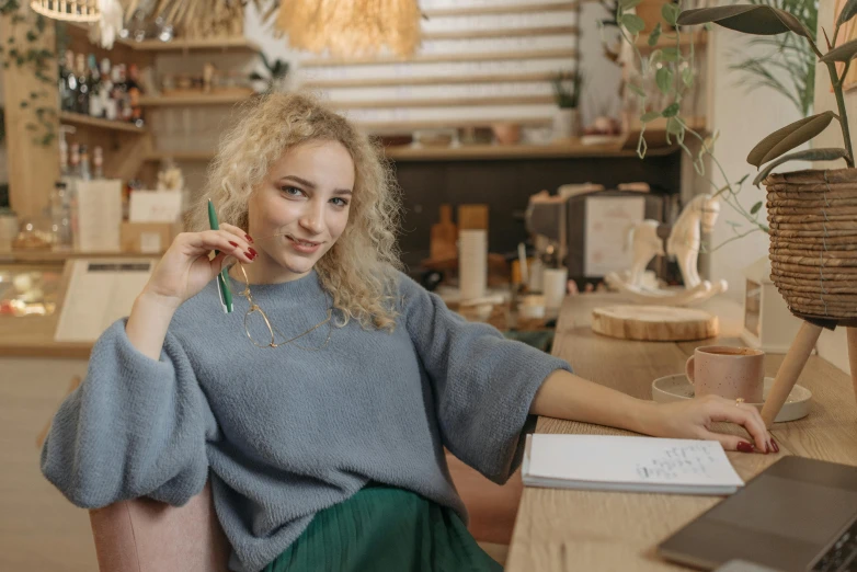 a woman sitting at a desk in front of a laptop, a portrait, trending on pexels, academic art, curly blond, wearing casual sweater, holding pencil, sitting in a lounge