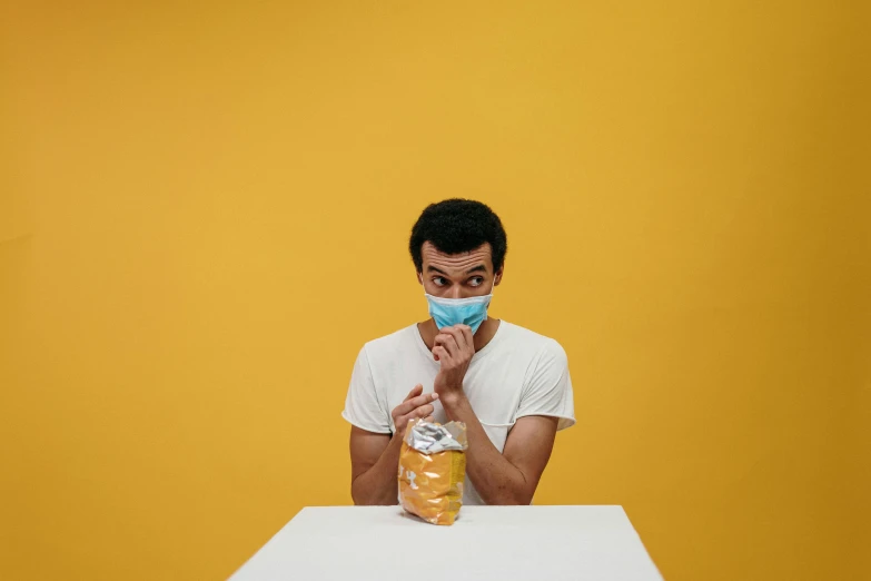 a man sitting at a table with a doughnut in front of him, by Adam Marczyński, pexels contest winner, surgical mask covering mouth, yellow backdrop, head in a jar, surprised