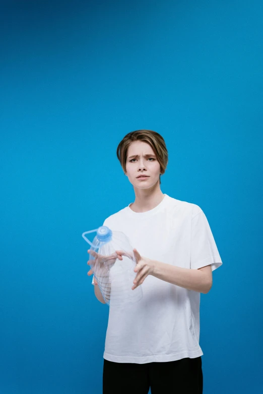 a man in a white shirt holding a water bottle, by Julia Pishtar, happening, cyan photographic backdrop, she's sad, tomboy, wearing a baggy