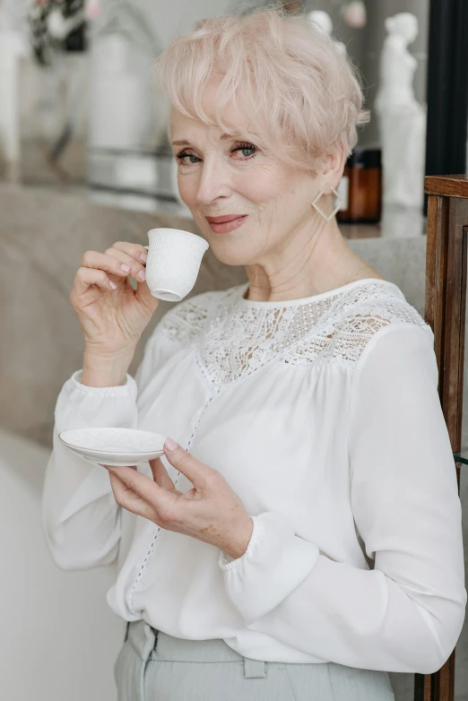 a woman holding a cup and saucer in her hands, inspired by Joanna Carrington, trending on pexels, rococo, with short bobbed white hair, a beautiful woman in white, age lines, with a drink